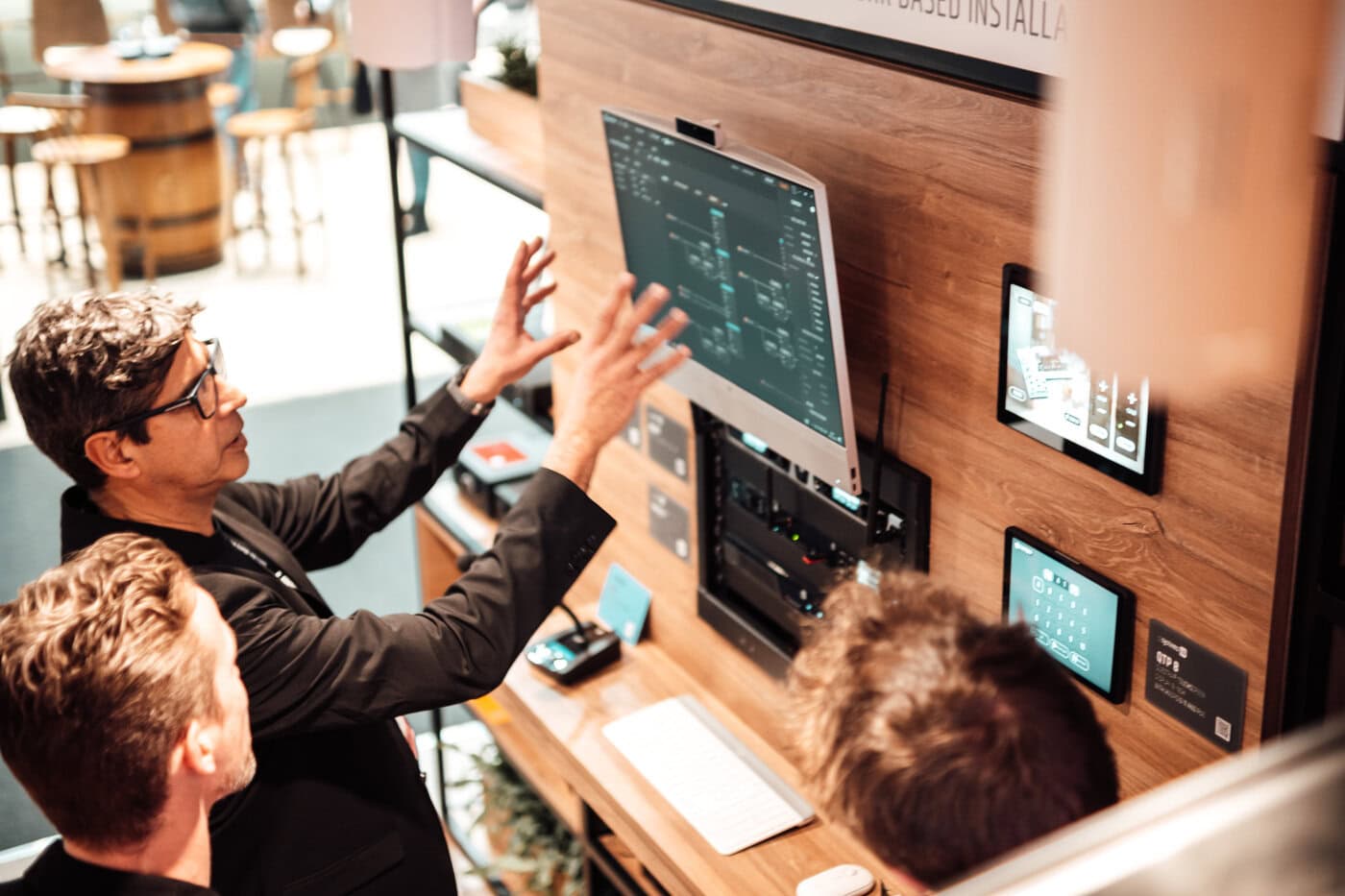 Zwei Personen stehen an einem Arbeitsplatz mit einer Holzwand und interagieren mit einem großen, an der Wand befestigten Touchscreen, auf dem technische Diagramme und mehrere kleinere Bildschirme angezeigt werden.