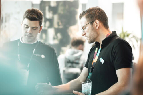 Zwei Männer mit schwarzen Hemden und Konferenzbändern stehen in einem Raum und unterhalten sich. Einer gestikuliert mit der Hand, während der andere aufmerksam zuhört.