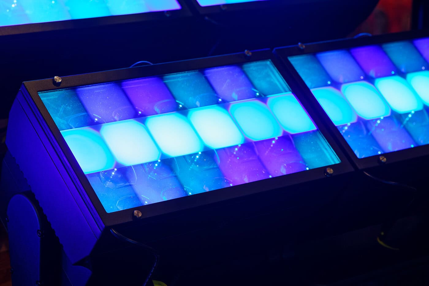 Nahaufnahme von rechteckigen LED-Bühnenscheinwerfern, die helle blaue, türkise und violette Farbtöne ausstrahlen und in einem schwarzen Metallgehäuse gitterförmig angeordnet sind.