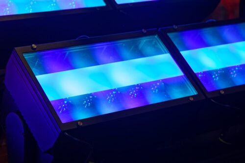 Nahaufnahme von zwei rechteckigen LED-Bühnenscheinwerfern, die blaues und violettes Licht ausstrahlen, mit sichtbaren Reflexionen auf ihren Glasflächen.