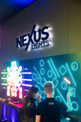 Zwei Personen interagieren mit einem Wanddisplay aus hell erleuchteten geometrischen Formen unter einem NEXUS LIGHTS-Schild in einer Ausstellung oder einem Ausstellungsraum.