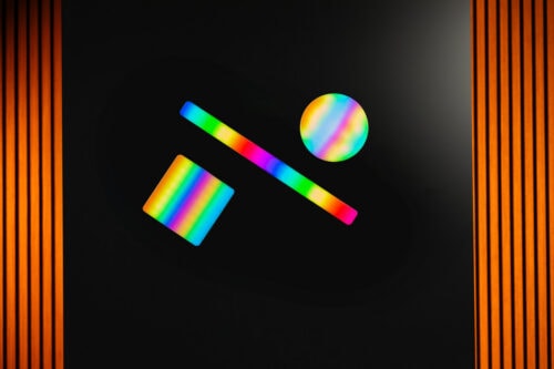 Auf einem schwarzen Hintergrund mit orangefarbenen vertikalen Streifen an den Seiten befinden sich drei geometrische Formen in Regenbogenfarben: ein gekipptes Rechteck, eine horizontale Linie und ein Kreis.