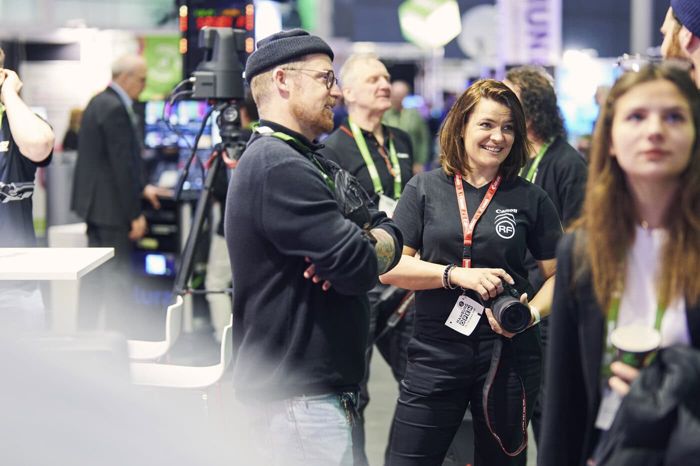 OP25_AW_150125_503 Eine Gruppe von Menschen steht und unterhält sich bei einer Indoor-Veranstaltung; eine Frau mit einer Kamera lächelt, während andere zuhören oder sich umschauen.