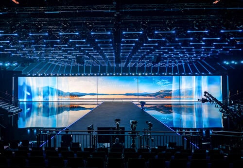 Eine große, leere Bühne mit einem großen digitalen Bildschirm, auf dem eine Landschaft mit Bergen und einem See zu sehen ist, umgeben von blauer Beleuchtung und Filmausrüstung im Vordergrund.