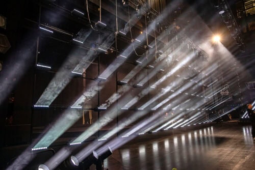 Mehrere Reihen von Bühnenscheinwerfern leuchten mit hellen Strahlen über eine dunkle Theaterbühne, wobei leichter Dunst die Strahlen durch die Luft sichtbar macht.