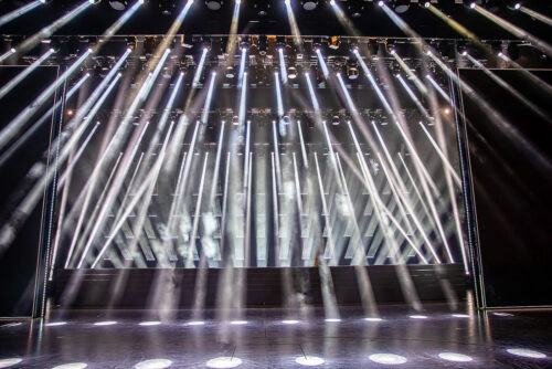 Breite Bühne mit zahlreichen weißen Scheinwerfern, die in einem Winkel auf den Boden gerichtet sind und in einem leeren Theater helle Strahlen und Dunst erzeugen.