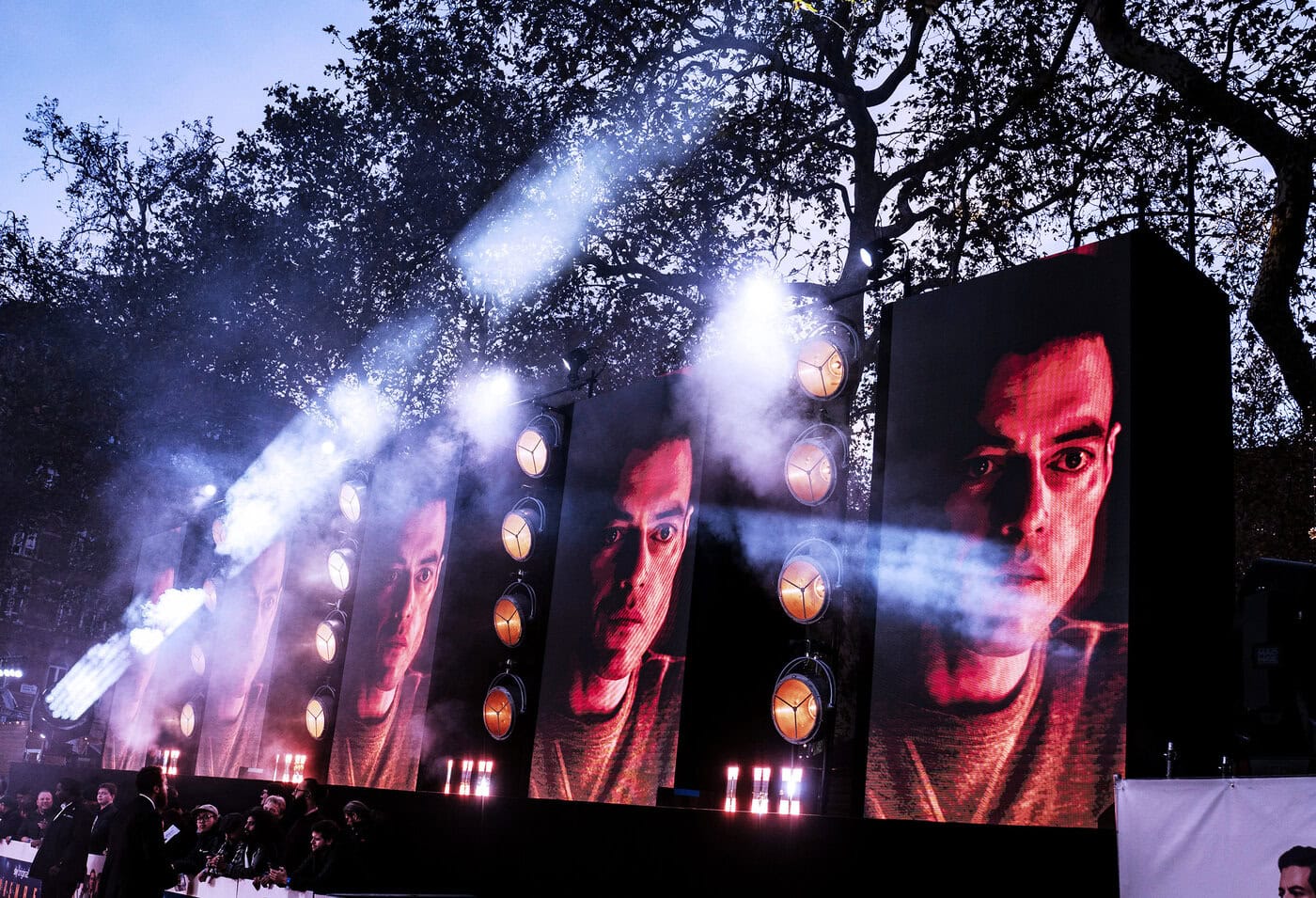 Große digitale Bildschirme im Freien zeigen das Gesicht eines Mannes, mit hellen Scheinwerfern und Nebeleffekten, und im Hintergrund sind Äste zu sehen.