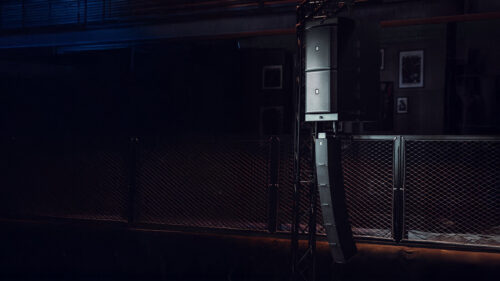 Ein vertikales Line-Array-Lautsprechersystem ist an einer Metalltraverse neben einem schwarzen Maschendrahtzaun in einer schwach beleuchteten Halle montiert.