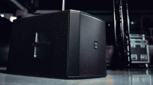 Ein schwarzer professioneller Audio-Lautsprecher mit einer Netzfront steht auf einem reflektierenden Boden in einem Innenraum, mit Gerätekisten und Strukturen im Hintergrund.