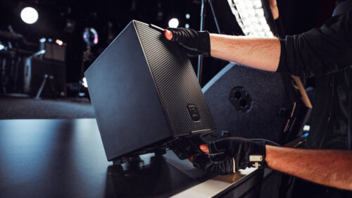 Eine Person mit Handschuhen platziert oder justiert einen schwarzen Lautsprecher auf einer Oberfläche in einer Bühnen- oder Studioumgebung mit audiovisuellen Geräten im Hintergrund.