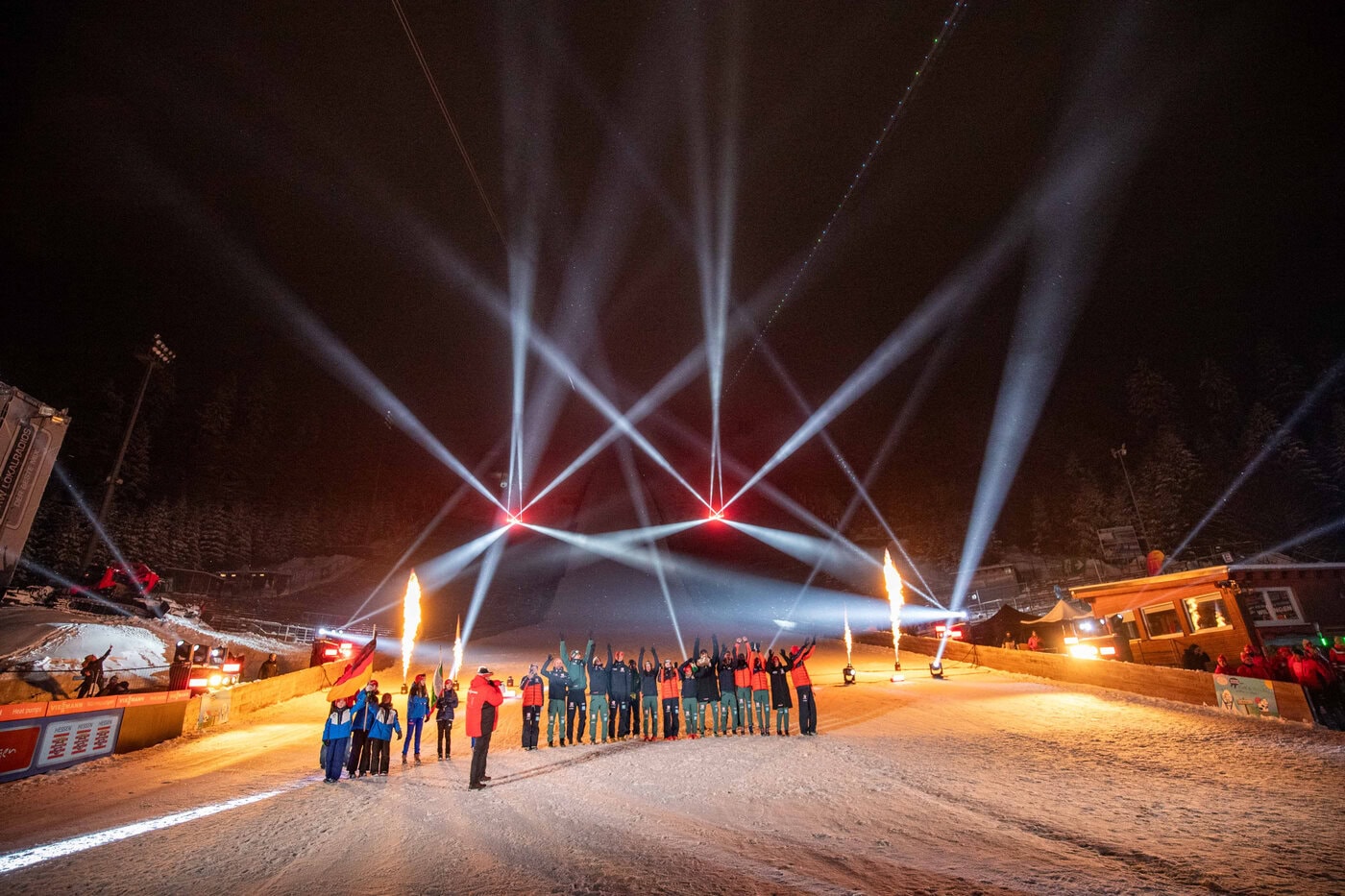 Cameo beim Skisprung-Weltcup Eine Gruppe von Menschen steht nachts auf einem schneebedeckten Hang, während im Hintergrund Scheinwerfer und Flammen eine festliche Atmosphäre schaffen.