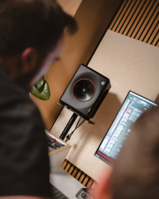 Zwei Personen arbeiten an einem Computer mit Audiobearbeitungssoftware in der Nähe eines schwarzen Studiomonitors, der in einem modernen Aufnahmestudio an der Wand befestigt ist.