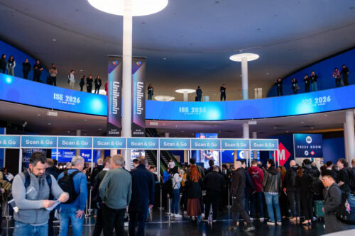 Eine große Gruppe von Menschen wartet in der Schlange vor einem Scan & Go-Registrierungsbereich für die ISE 2024 in einem modernen Gebäude mit blauer Beschilderung und einem kreisförmigen Balkon darüber.