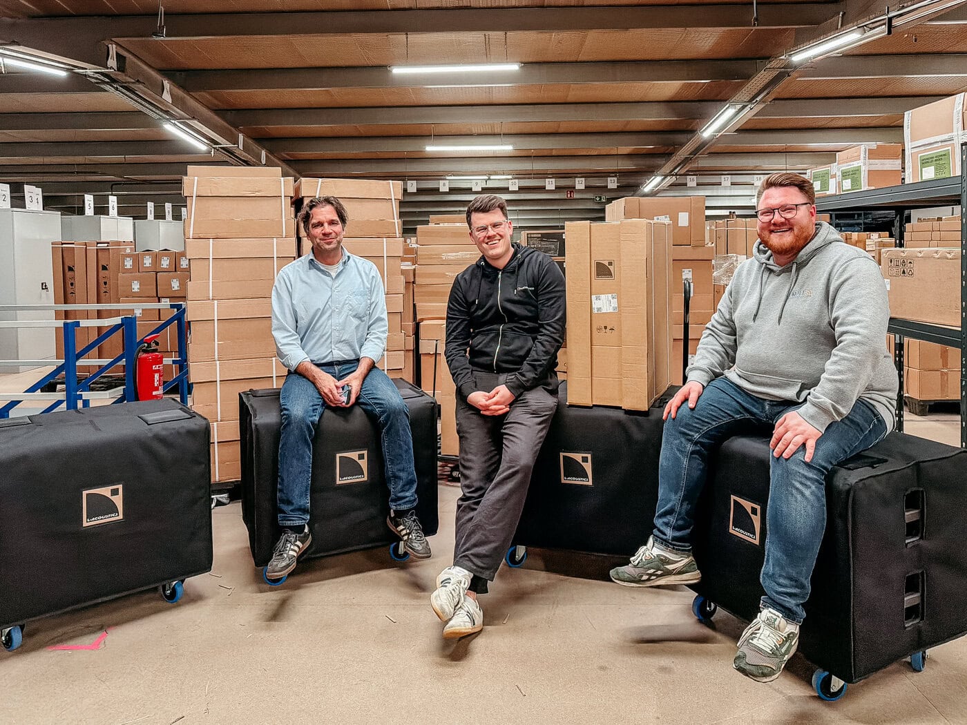 Drei Männer sitzen auf großen schwarzen Kisten mit Rädern in einer Lagerhalle, umgeben von gestapelten Kartons und Regalen, unter Neonlicht.