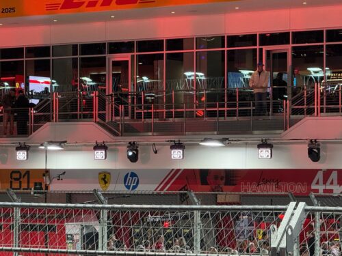 Ein Blick auf ein Formel-1-Fahrerlager mit Ferrari- und HP-Logos, großen Fenstern darüber und zwei Personen, die auf einem Balkon stehen und die Szene überblicken.