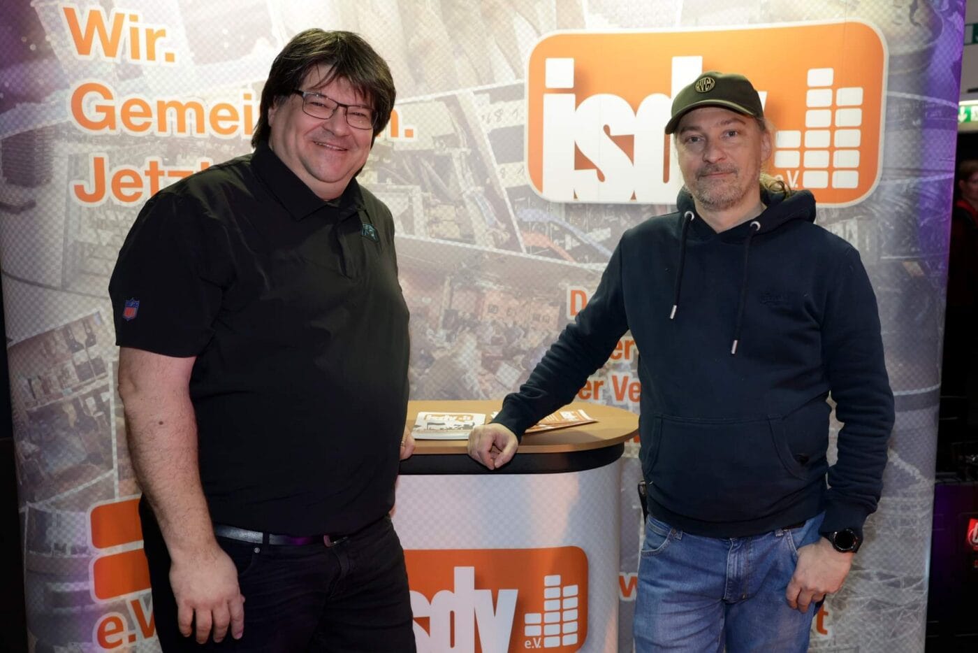 Zwei Männer stehen vor einem Stand mit einem Markenhintergrund, auf dem der Text ispy und deutsche Wörter zu lesen sind. Beide sind leger gekleidet und lächeln in die Kamera.