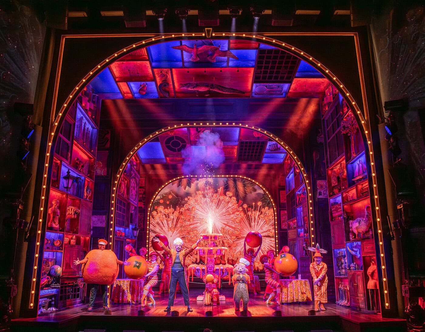 Schauspieler in farbenfrohen Kostümen, darunter auch überdimensionale Früchte, treten auf einer hell erleuchteten, aufwendig gestalteten Bühne auf, die mit einem Feuerwerk und farbenfrohen Dekorationen im Hintergrund geschmückt ist.