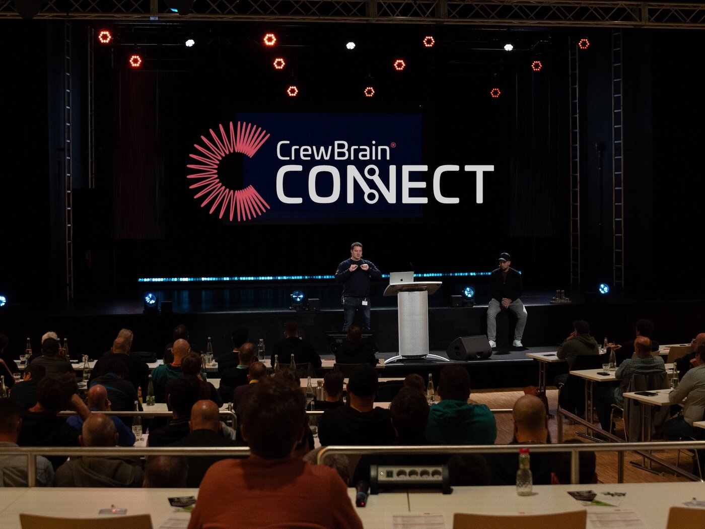 Ein Redner steht auf der Bühne neben einer sitzenden Person vor dem Publikum, hinter der ein großes CrewBrain CONNECT-Schild angebracht ist.