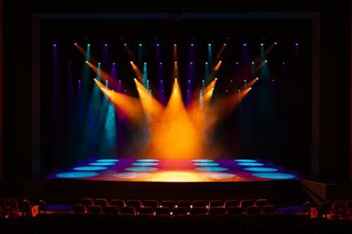 Eine leere Theaterbühne ist mit orangefarbenen, blauen und violetten Scheinwerfern beleuchtet, die farbige Muster auf dem Boden erzeugen. Die Sitze im Zuschauerraum sind unbesetzt.