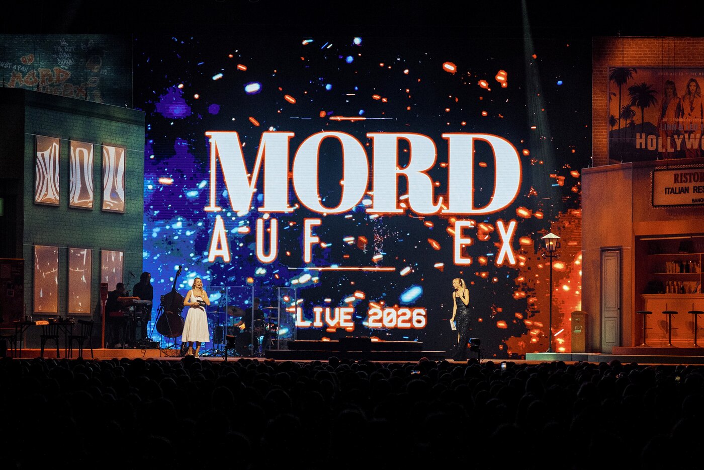 Zwei Personen stehen auf der Bühne vor einem großen Bildschirm, auf dem MORD AUF EX LIVE 2026 zu sehen ist, mit bunten Lichtern und Kulissen, die an Gebäude auf beiden Seiten erinnern.