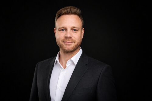 Ein Mann mit kurzen hellbraunen Haaren und einem Bart trägt ein weißes Hemd und einen dunklen Blazer und posiert vor einem schlichten schwarzen Hintergrund.