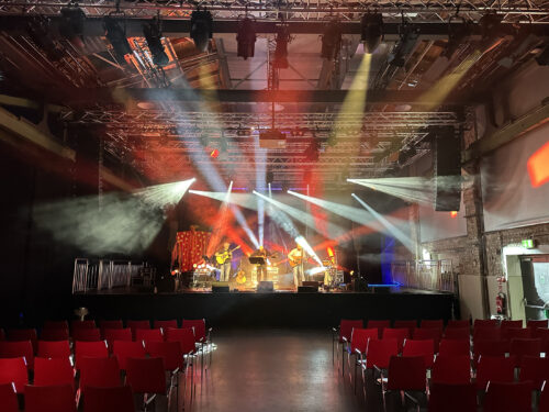 Eine Bühne mit vier Musikern, die unter buntem Bühnenlicht in einer Halle spielen; im Vordergrund sind leere Reihen roter Stühle zu sehen.