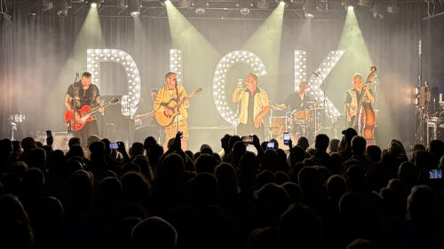Eine Band spielt auf der Bühne, im Hintergrund sind große Leuchtbuchstaben mit der Aufschrift DICK zu sehen, während die Menge zusieht und Fotos macht.