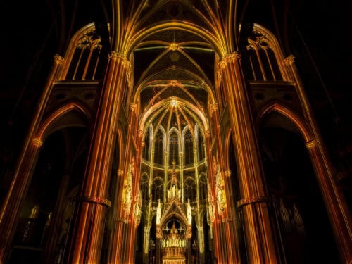 Das Innere einer Kathedrale mit gewölbten Decken, Bögen und Säulen wird durch goldene und orangefarbene Lichteffekte beleuchtet, die die architektonischen Details hervorheben.