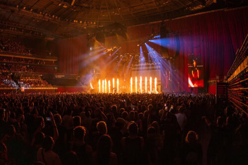 Ein großes Hallenkonzert mit einer Menschenmenge, die vor einer Bühne steht, die von orangefarbenen Flammen und bunten Lichtern erleuchtet wird, wobei im Hintergrund Vorhänge und Sitzgelegenheiten zu sehen sind.