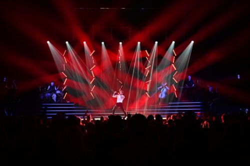 Eine Band spielt auf der Bühne mit dramatischen roten und weißen Lichtern, die nach außen strahlen, während die Menge im Vordergrund zusieht.