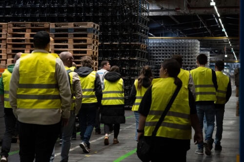 Eine Gruppe von Menschen in gelben Warnwesten geht unter industrieller Beleuchtung durch ein Lagerhaus mit gestapelten Paletten und Metallregalen.