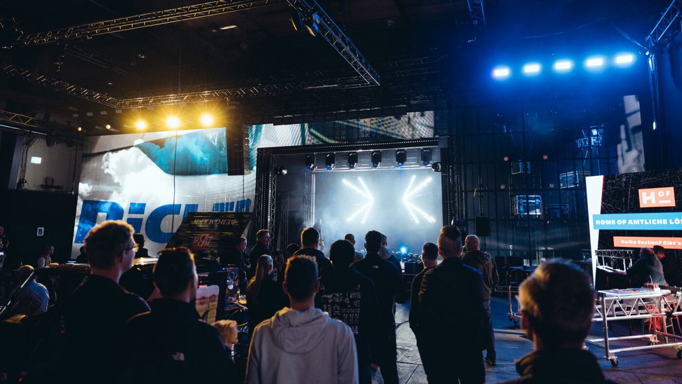 Bühnenshow bei den NicLen Family Open House Days 2026 Eine Gruppe von Menschen steht in einem dunklen Veranstaltungsraum im Industrial-Stil und blickt auf eine hell erleuchtete Bühne mit Scheinwerfern und großen digitalen Bildschirmen.