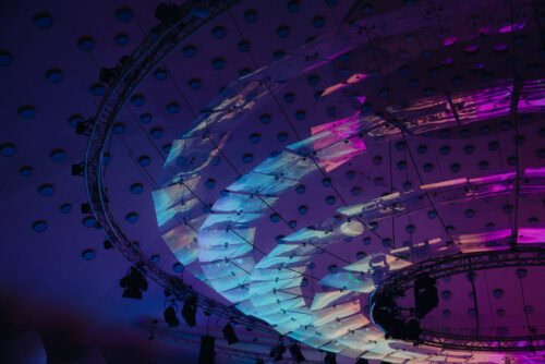 Deckeninstallation mit sich überlagernden, lichtdurchlässigen Paneelen, die von violetten und blauen Lichtern beleuchtet werden, vor einer Kuppel mit kreisförmigen Mustern und Metallrahmen.