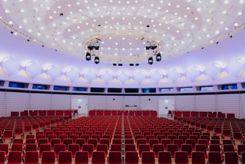 Großes, modernes Auditorium mit roten Sitzreihen vor einer Bühne. Die Decke ist mit kreisförmiger Beleuchtung und geometrischem weißen Dekor versehen. Der Raum ist leer.