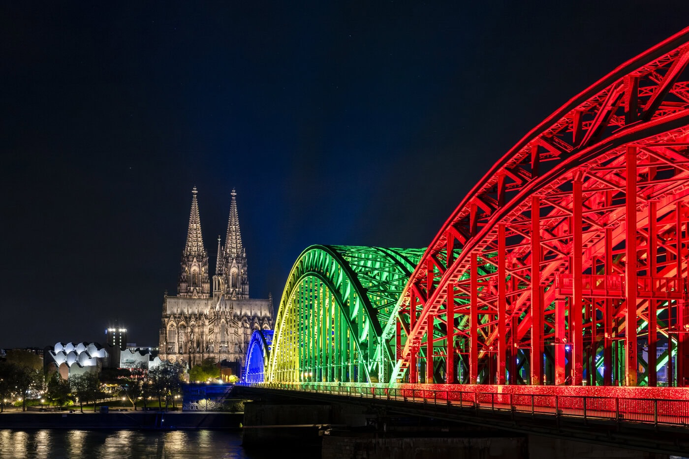 Die Hohenzollernbrücke in Köln ist nachts in Regenbogenfarben beleuchtet, im Hintergrund ist der beleuchtete Kölner Dom zu sehen.