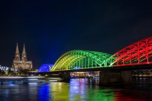 Eine in Regenbogenfarben beleuchtete Brücke überspannt nachts einen Fluss, im Hintergrund sind eine große Kathedrale und die Lichter der Stadt zu sehen.