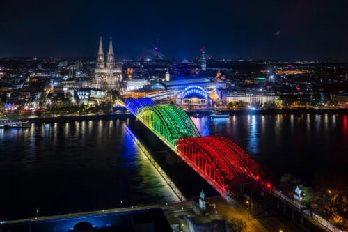 Eine Nachtansicht einer Stadt mit einer großen, in Regenbogenfarben beleuchteten Brücke über einem Fluss sowie einer Kathedrale und beleuchteten Gebäuden im Hintergrund.