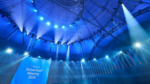 Ein großer, moderner Saal mit blauer Beleuchtung, Scheinwerfern an der Decke und einer Leinwand, auf der „Global Staff Meeting 2026“ zu sehen ist.