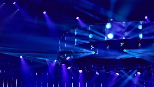 Eine kreisförmige Bühnenbeleuchtung mit mehreren blauen Scheinwerfern und Lichtstrahlen, die in einem dunklen Saal in verschiedene Richtungen leuchten.