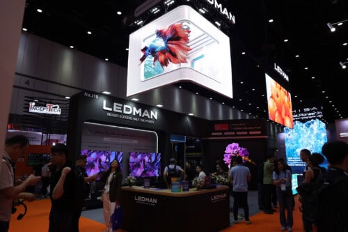 Ein Messestand mit der Aufschrift „LEDMAN“, großen LED-Bildschirmen, auf denen farbenfrohe Bilder zu sehen sind, und Menschen, die auf einem orangefarbenen Teppichboden umhergehen und miteinander interagieren.