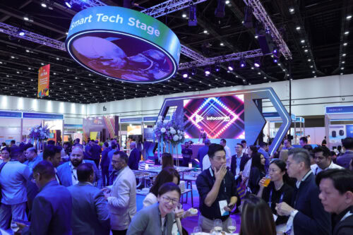 Die Besucher versammeln sich bei einer Tech-Veranstaltung in den Innenräumen, bei der es Stände, Essen und Getränke gibt. Im Hintergrund sind ein großes „Smart Tech Stage“-Schild und ein digitaler Bildschirm zu sehen.