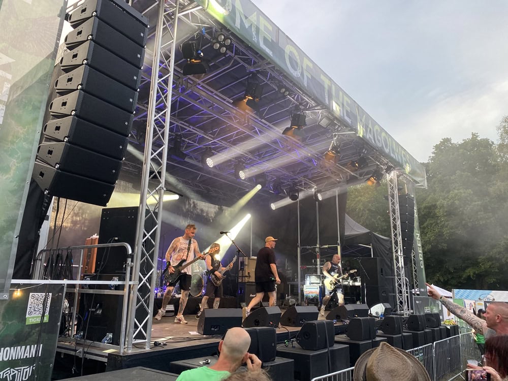 Eine Band tritt auf einer Freiluftbühne mit Beleuchtung und Lautsprechern auf, während eine Menschenmenge zusieht. Im Hintergrund sind auf der rechten Seite des Bildes Bäume zu sehen.