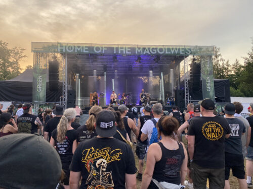 Eine Menschenmenge verfolgt den Auftritt einer Live-Band auf einer Außenbühne bei einem Musikfestival. Auf dem Banner über der Bühne steht „HOME OF THE MAGOLWES“. Im Hintergrund sind Bäume und der Himmel zu sehen.