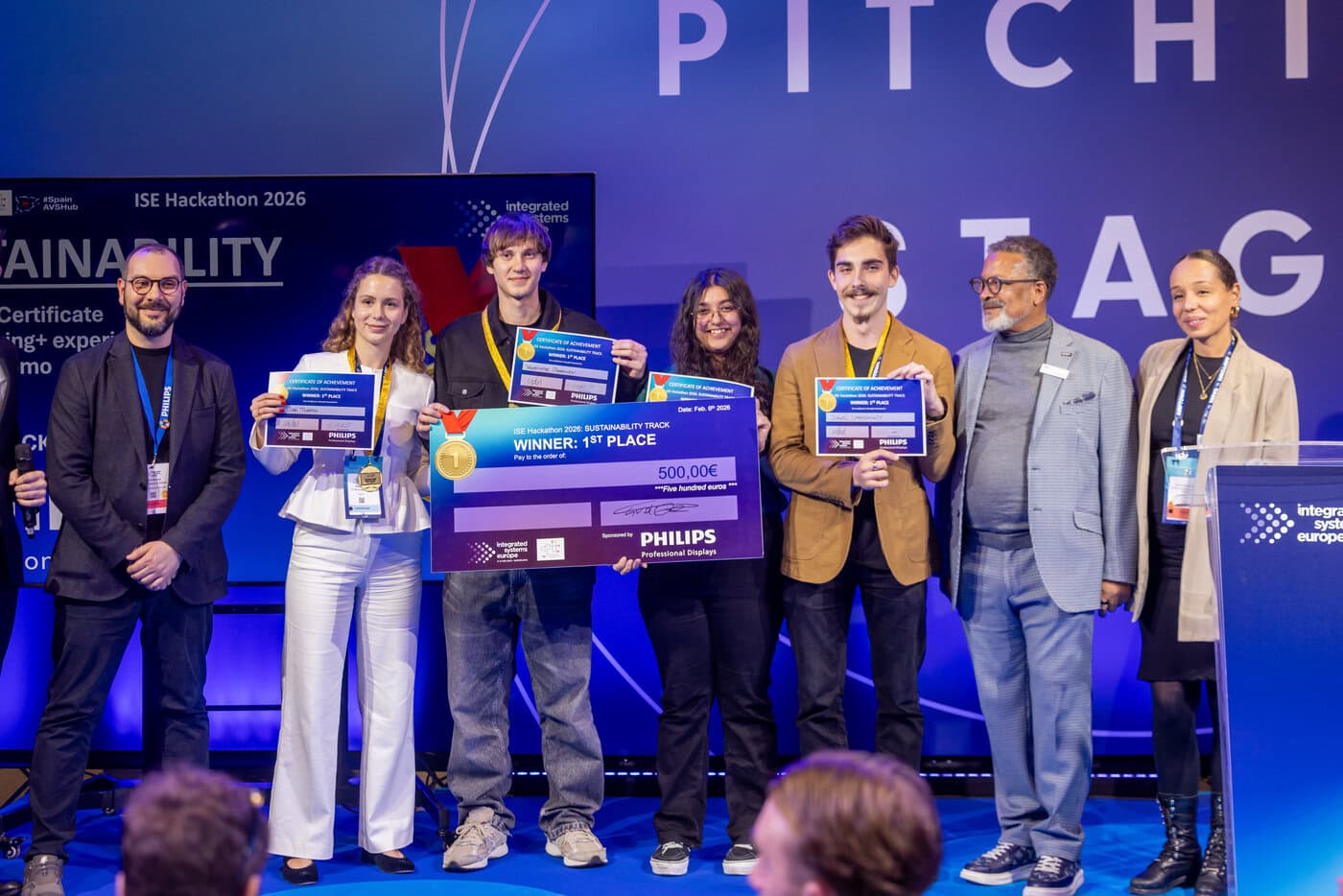 Day 4 – 6 Feb_4 3 Stages_4 3 4 Pitching Stage – Hackathon Awards Ceremony – CS300_Hackathon Final Awards Ceremony_475A8597 JPG_8192x5464 Eine Gruppe von sechs Personen steht auf der Bühne und hält einen großen Scheck sowie Urkunden in den Händen, während zwei weitere Personen neben ihnen stehen. Im Hintergrund sind die Beschilderung und das Logo der Veranstaltung zu sehen.