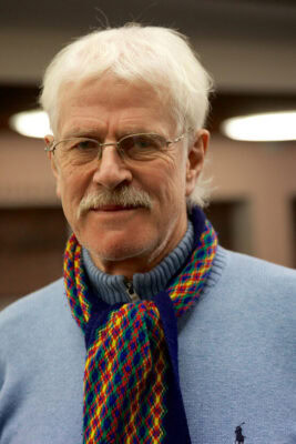 Ein älterer Mann mit weißem Haar, Brille und Schnurrbart, der einen blauen Pullover und einen bunten Schal trägt und in einem Innenraum mit unscharfen Lichtern im Hintergrund in die Kamera blickt.