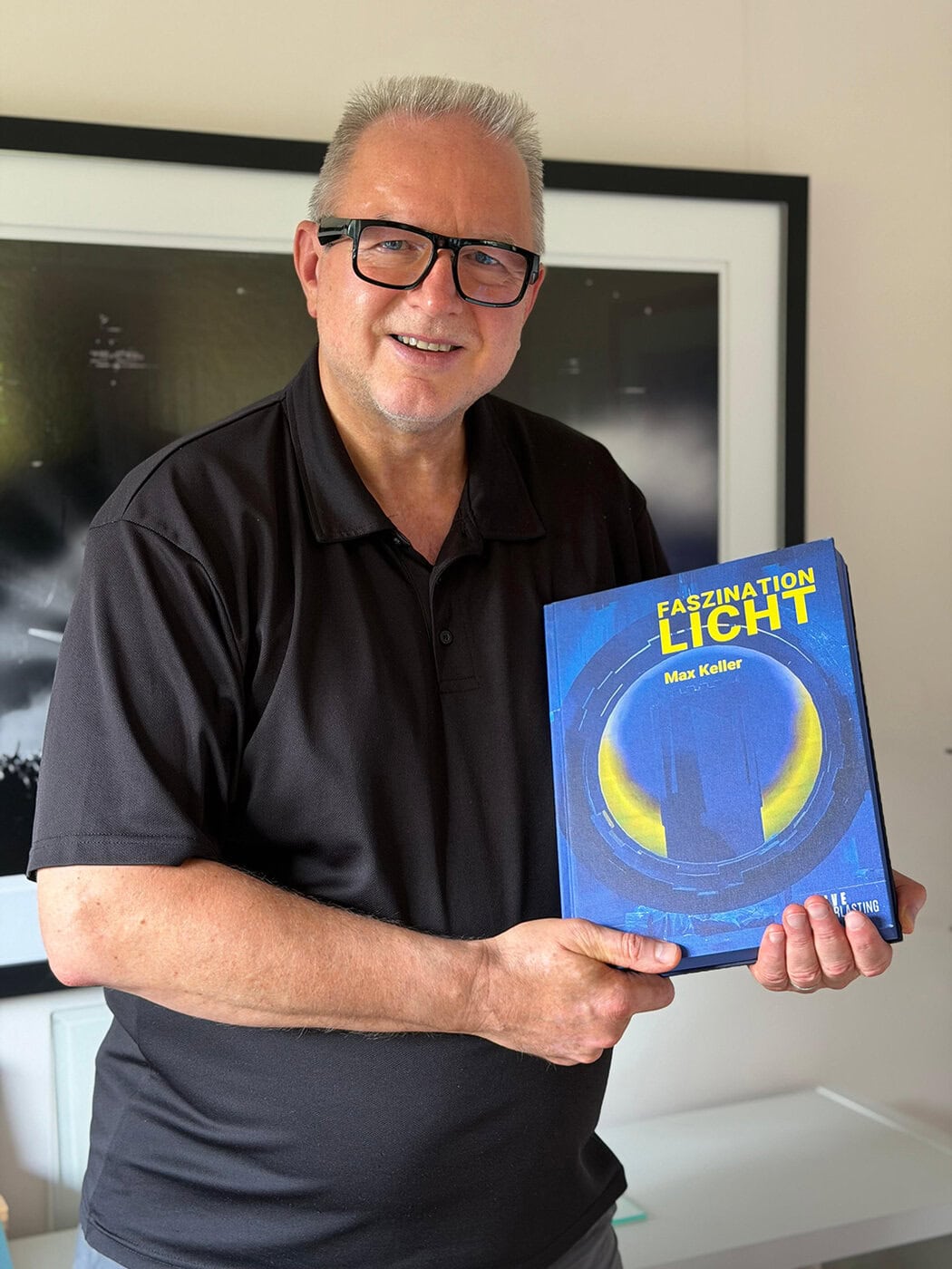 Faszination-Licht-03-Ralph Larmann-kein Copright Ein Mann mit Brille und in einem schwarzen Poloshirt hält ein Buch mit dem Titel „Faszination Licht“ von Max Keller in der Hand und steht in einem Raum vor gerahmten Kunstwerken.