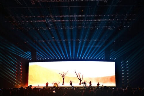 Eine Band tritt auf der Bühne vor einer großen Leinwand auf, auf der eine Wüstenlandschaft zu sehen ist, während blaues Bühnenlicht über einer Menschenmenge nach außen strahlt.
