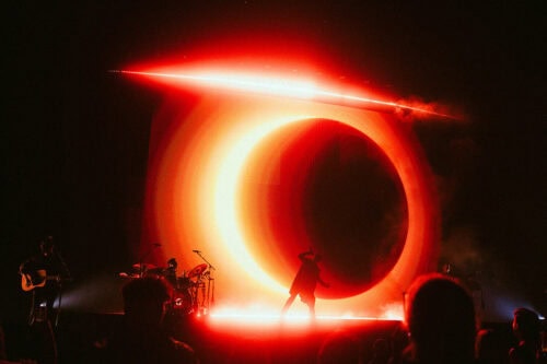 Eine Band tritt auf der Bühne auf, hinter ihr befindet sich eine große, leuchtend rote, kreisförmige Lichtinstallation, die für eine dramatische Beleuchtung sorgt und die Silhouetten der Musiker und des Publikums hervorhebt.