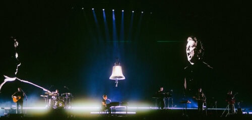 Eine Band tritt auf der Bühne auf, in deren Mitte ein Pianist steht; auf Leinwänden werden große Gesichter projiziert, über der Bühne hängt eine Glocke, und die dramatische Beleuchtung lässt die Instrumente erkennen.