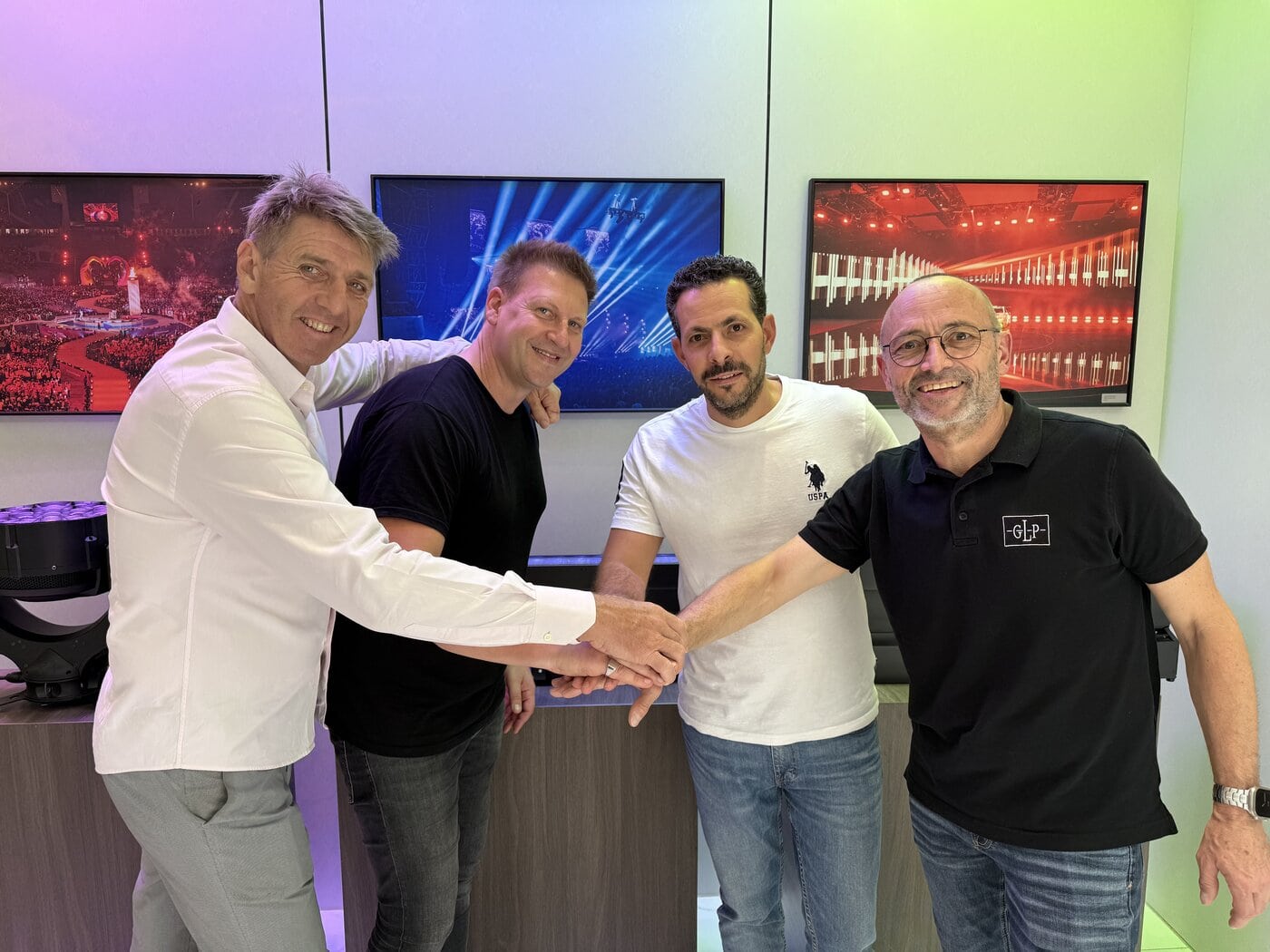 Left To Right_Søren Storm Davey Taylor Eli Malka Udo Künzler_002 Vier Männer stehen in einem Raum und geben sich in einer Gruppe die Hand. Hinter ihnen sind drei an der Wand befestigte Bildschirme zu sehen, auf denen Bilder von Konzerten oder Veranstaltungen mit Bühnenbeleuchtung zu sehen sind.